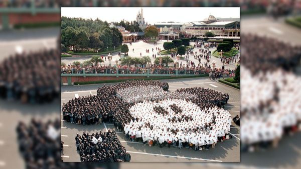 In a parking lot, people form Mickey Mouse's face. People surround the fence that divides the lot from the rest of Tokyo Disneyland's park, which you can see in the distance.