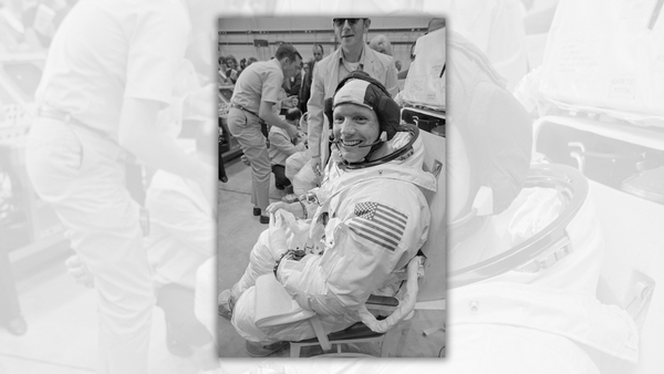 A white man smiles while wearing an astronaut suit. People stand behind him.
