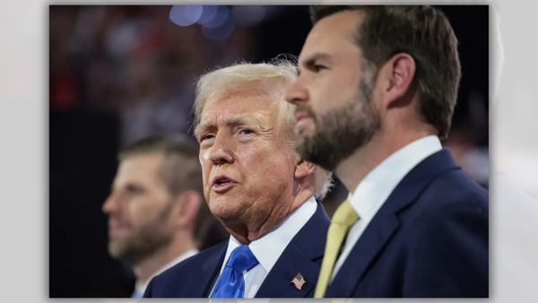 A white man wearing a blue suit stands next to another white man wearing a blue suit. A white person can be seen in the background of the image.